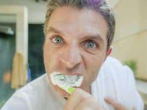 lifestyle portrait of young attractive white man with blue eyes at home bathroom brushing tooth with toothbrush energically looking at himself on toilet mirror in dental health care concept