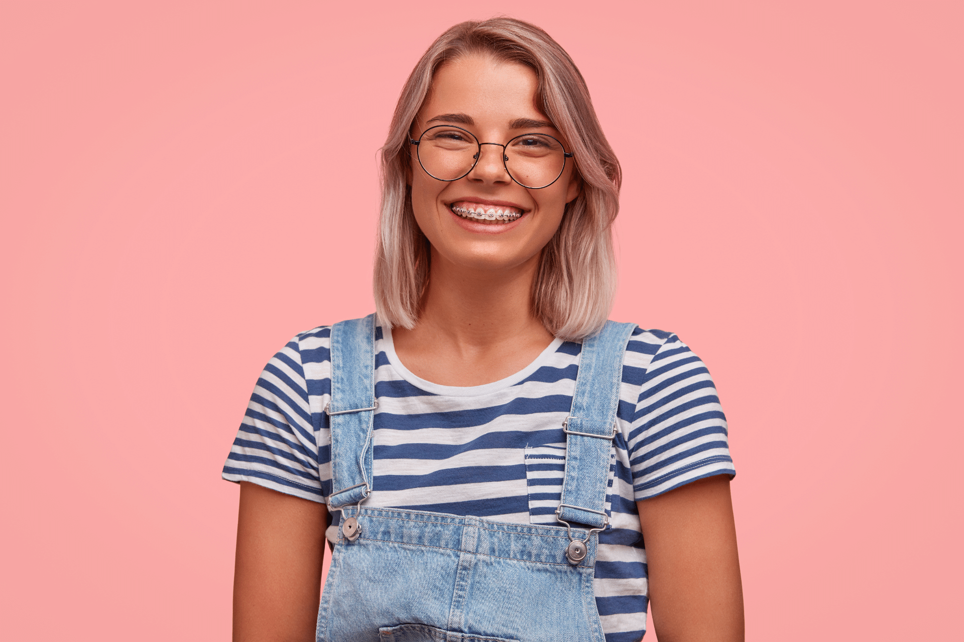Young woman wearing glasses and smiling with metal braces against a pink background.
